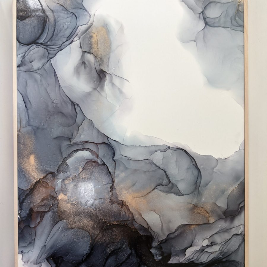 A dark and light blue abstract art piece, with spashes of gold, sits within a light wooden frame against a white background.  The blue stretches along the left-hand side of the piece, reaching up from the bottom.  The top right of the piece has a mark of light blue that reaches towards the side, but does not reach, creating a central area of whitespace.