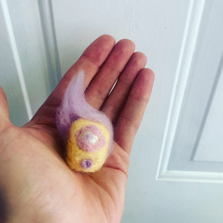 Needle felted yellow and purple cyclops surfer character being held in the palm of a hand.