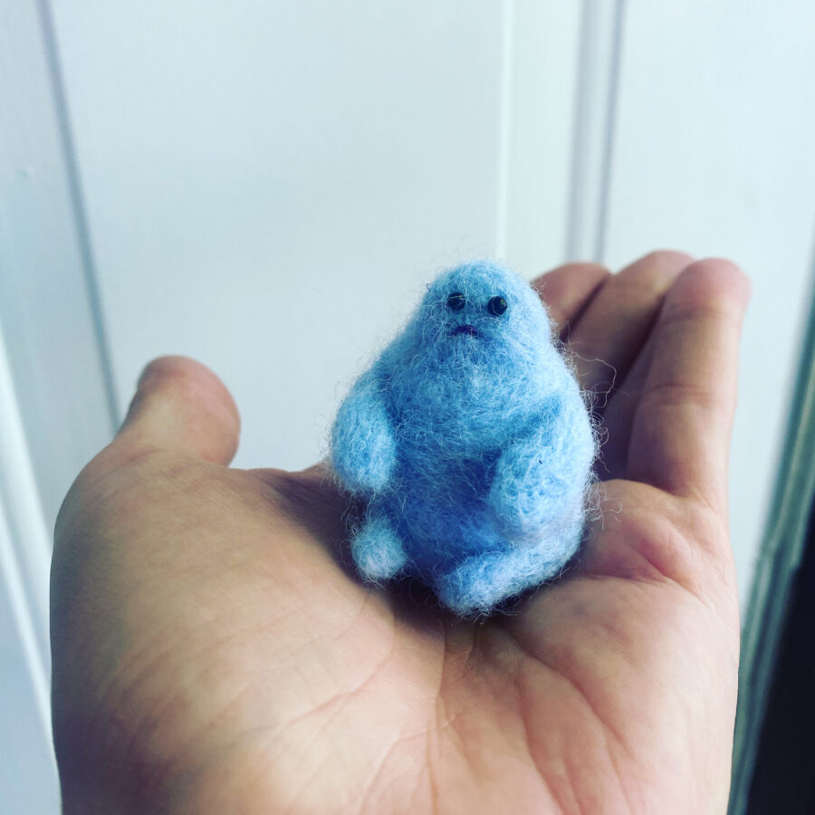Needle felted blue sad character being held in the palm of a hand.