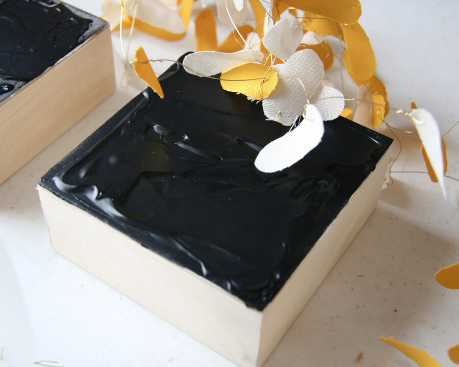 Image description: A detail image of thick black acrylic paint on a small square wood panel and small oval yellow and beige pieces of fabric hanging over from thin metal wire.