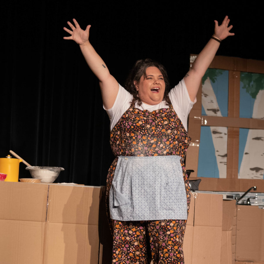 Carly performing in her critically-acclaimed solo show Meat(less) Loaf. She stands onstage in front of a cardboard kitchen arms stretched to the sky