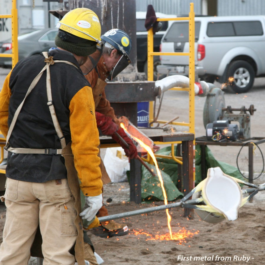 two persons attending first molten metal flow out of upright furnace outdoors