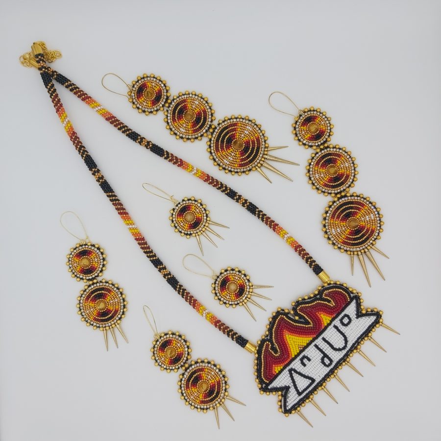 White background. Three pairs of earrings placed around a beaded medallion that has fire and Cree syllabics on it. All items are fire colours.