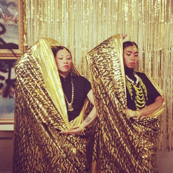 Robin Lacambra and Jen Maramba are in gold and black malongs painted by Mangopeeler, standing in front of a wall lined with gold tinsel, wearing lots of accessories and performing precolonial Filipino royalty.