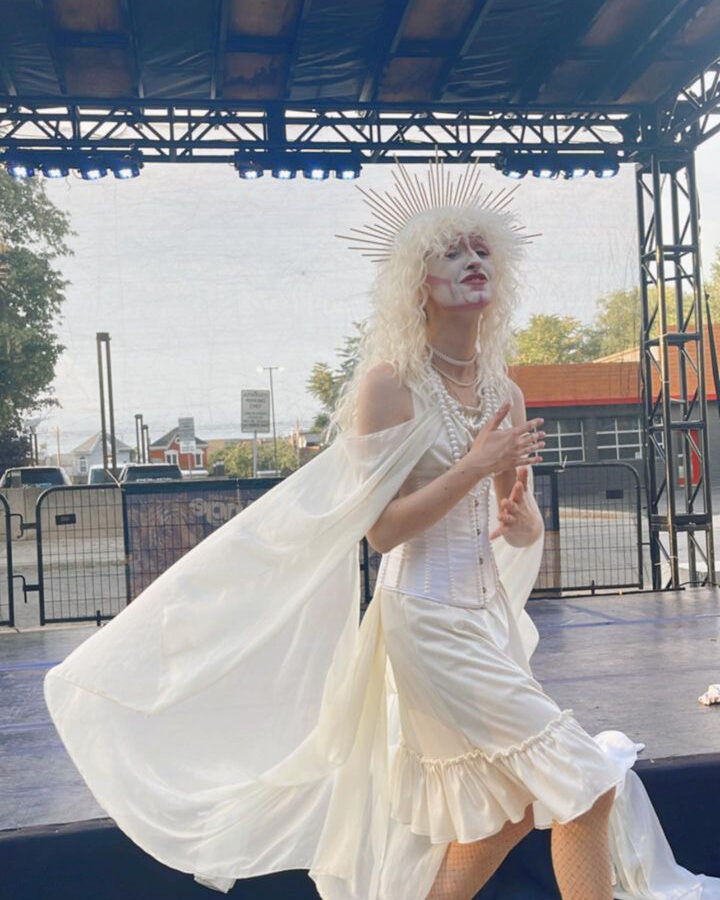 St. John Boy in his ghost look wearing all white with a painted white face and gold shoes. His white chiffon flows in the wind as he stands on the outdoor stage. At Hamilton Fringe Festival 2023.