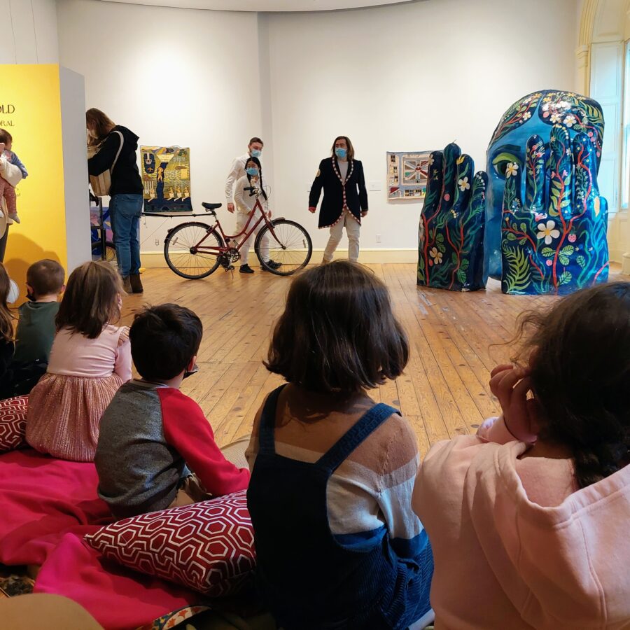 A group of children in WAHC's main gallery, viewed from behind, watching a colourful performance by Clay and Paper Theatre.