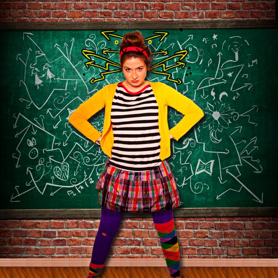 The ADHD Project promo image. A girl with brown hair in a messy bun, a black and white shirt, a tutu and miss-matched socks with red converse stares down the camera with their hands on their hips. The background is a green chalk board covered in doodles in chalk. Stick figure pictures and arrows pointing in all different directions coming from the head on the person. Brick lies behind the chalk board filling out the rest of the background