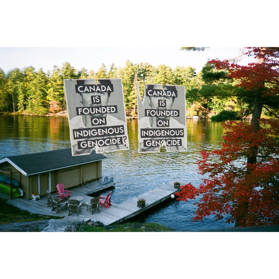 Photo shows the edge of a lake during a sunny day. The horizon is filled with trees and there is a tree on the right side, closer to the viewer that’s leaves are turning red. There is a dock and muskoka chairs backing onto the lake. In the middle of this, is 2 ripped posts of Justin Trudeau with the words “Canada is founded on Indigenous Genocide” on top.