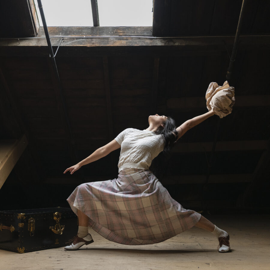 This is a photo from 'Enemy Lines'. Mayumi is dressed in 1940s skirt and knit top. She reaches back in a deep lunge with a ball of fabric in her upper hand. She is in front of a chest in a wood attic space.