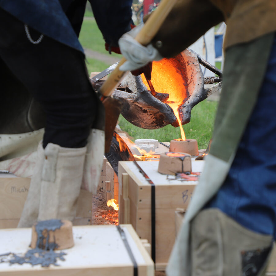 Close up of iron being poured from ladle into mould between legs of people in protective gear